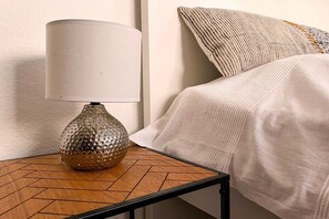 Uma mesa de cabeceira com abajur e um vaso texturizado, uma cama com cobertura branca e almofada estampada.