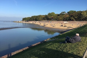 Uma praia com uma pessoa sentada na grama, mar calmo e árvores ao fundo.