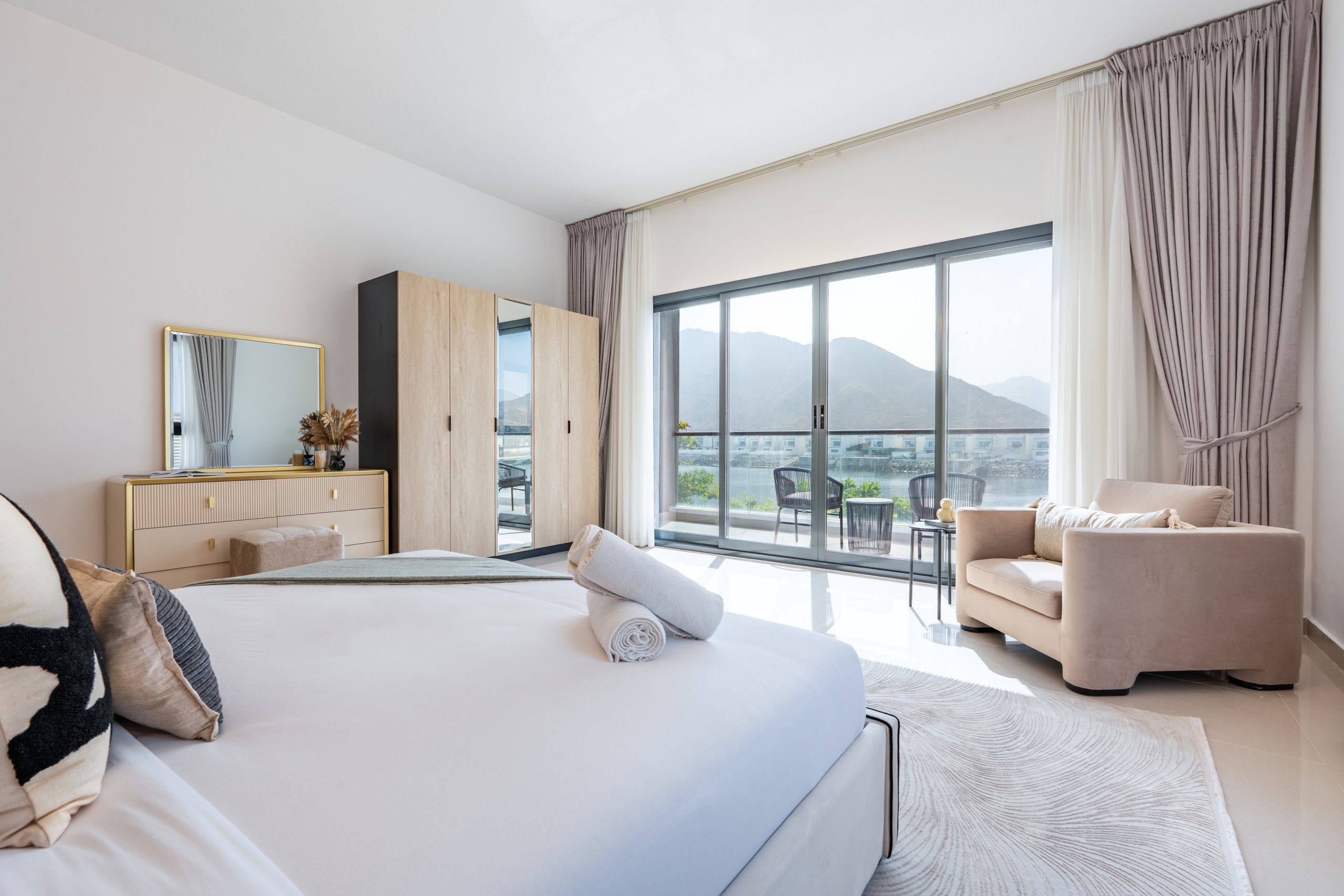 A modern hotel room with a large bed, a sitting area with a sofa and armchairs, a wooden wardrobe, and a balcony with a mountain view.