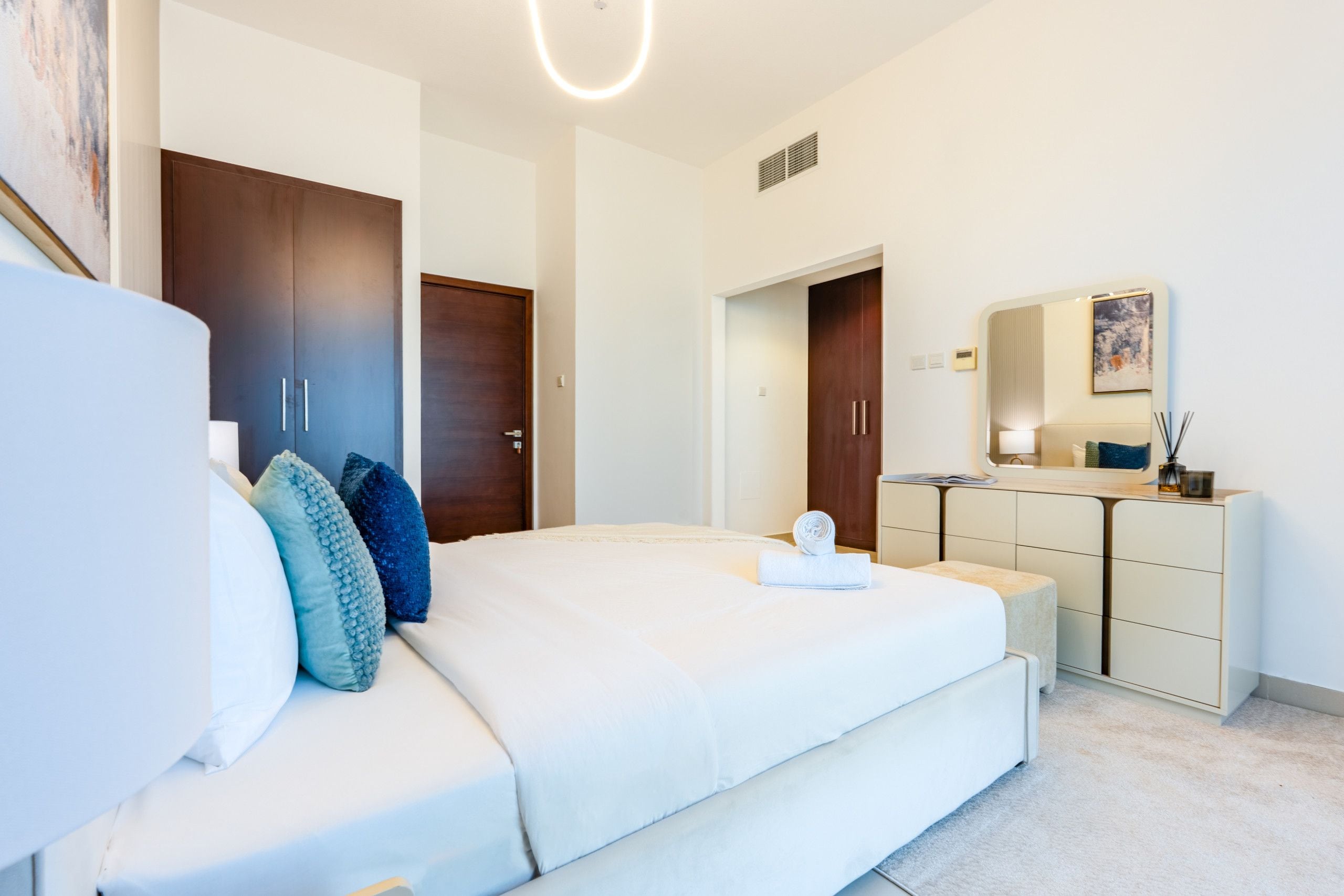 A modern bedroom with a large bed, a dresser, a mirror, and a door leading to another room.