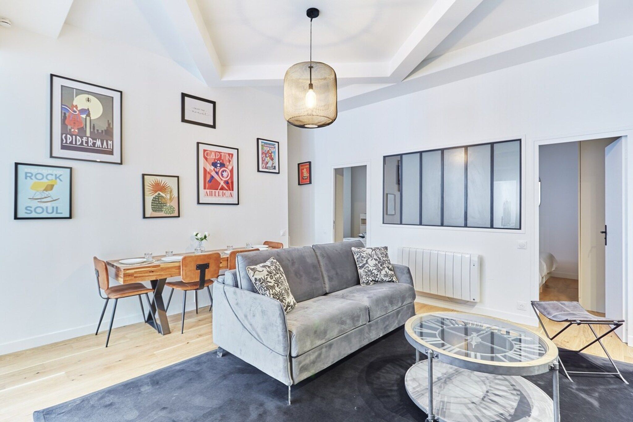 A modern living room with a grey sofa, a dining table with chairs, and a round coffee table. There are framed posters on the wall and a large window with blinds.