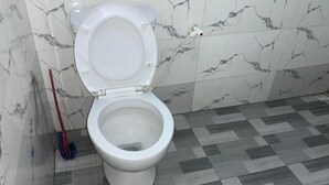 A toilet with a closed lid, a mop leaning against the wall, and a tiled floor with a geometric pattern. - OPTIZEE HOTEL & SUITES (Ikeja)