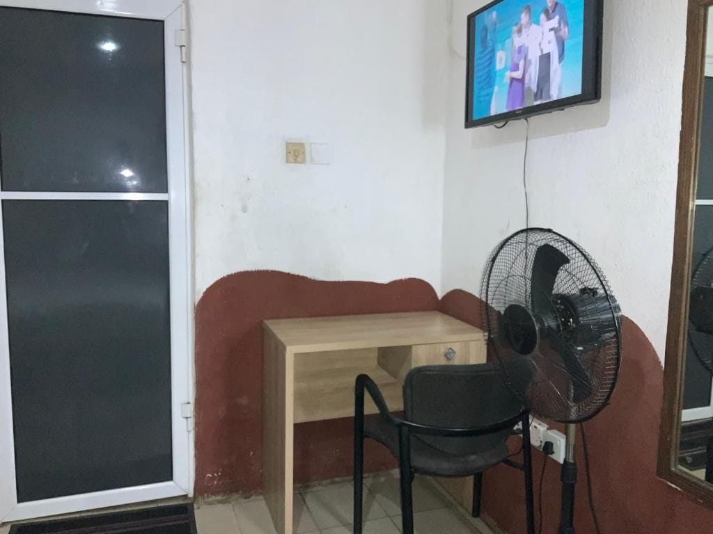 Een kamer met een houten bureau, een zwarte stoel, een ventilator en een aan de muur bevestigde televisie.