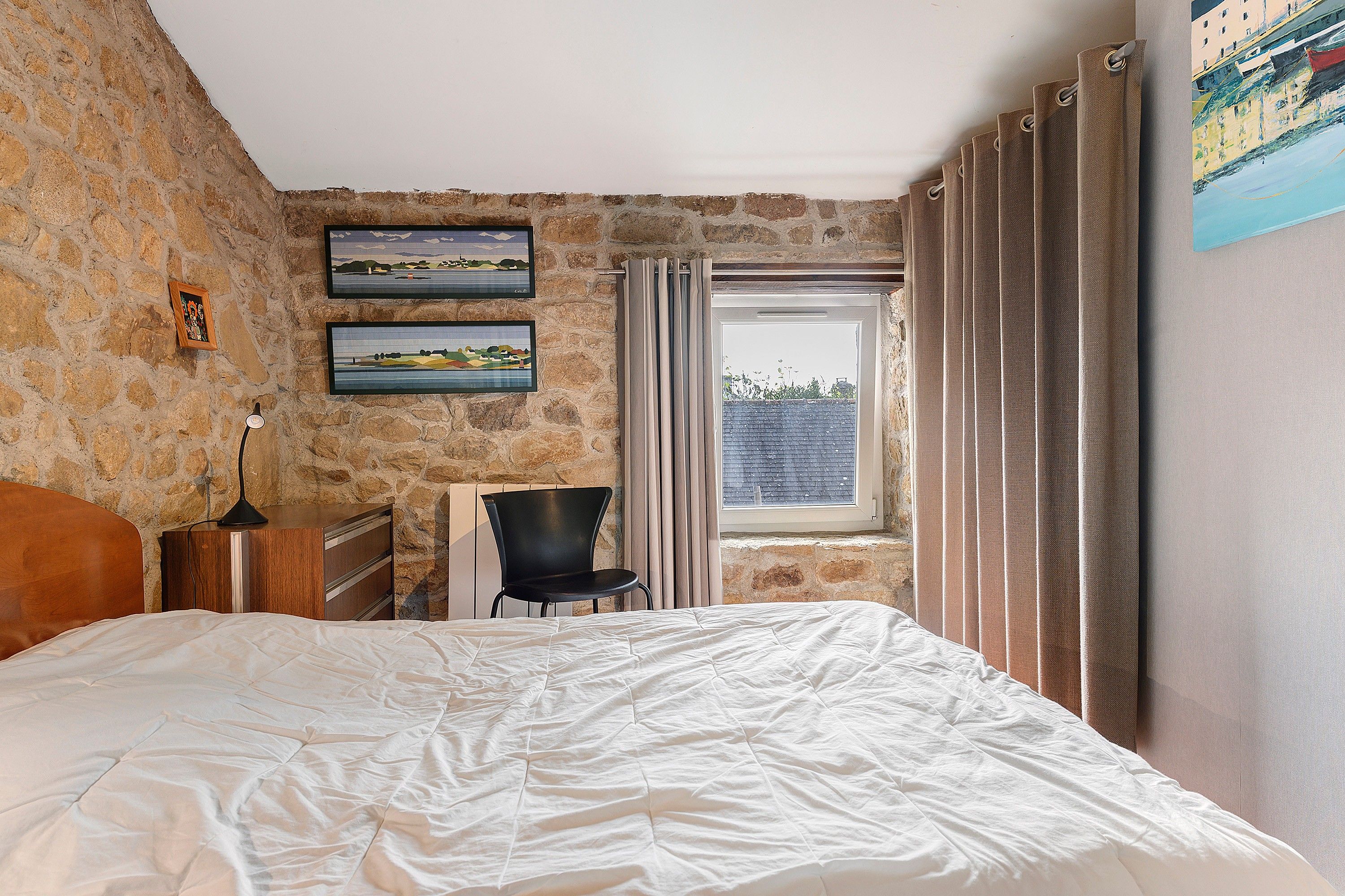 A bedroom with a stone wall, a bed, a chair, a desk, and a window with curtains.
