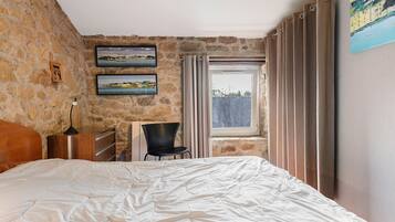 A bedroom with a stone wall, a bed, a chair, a desk, and a window with curtains.