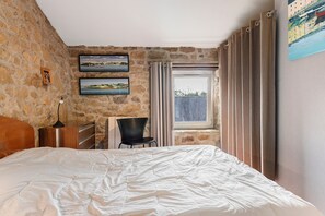 A bedroom with a stone wall, a bed, a chair, a desk, and a window with curtains.