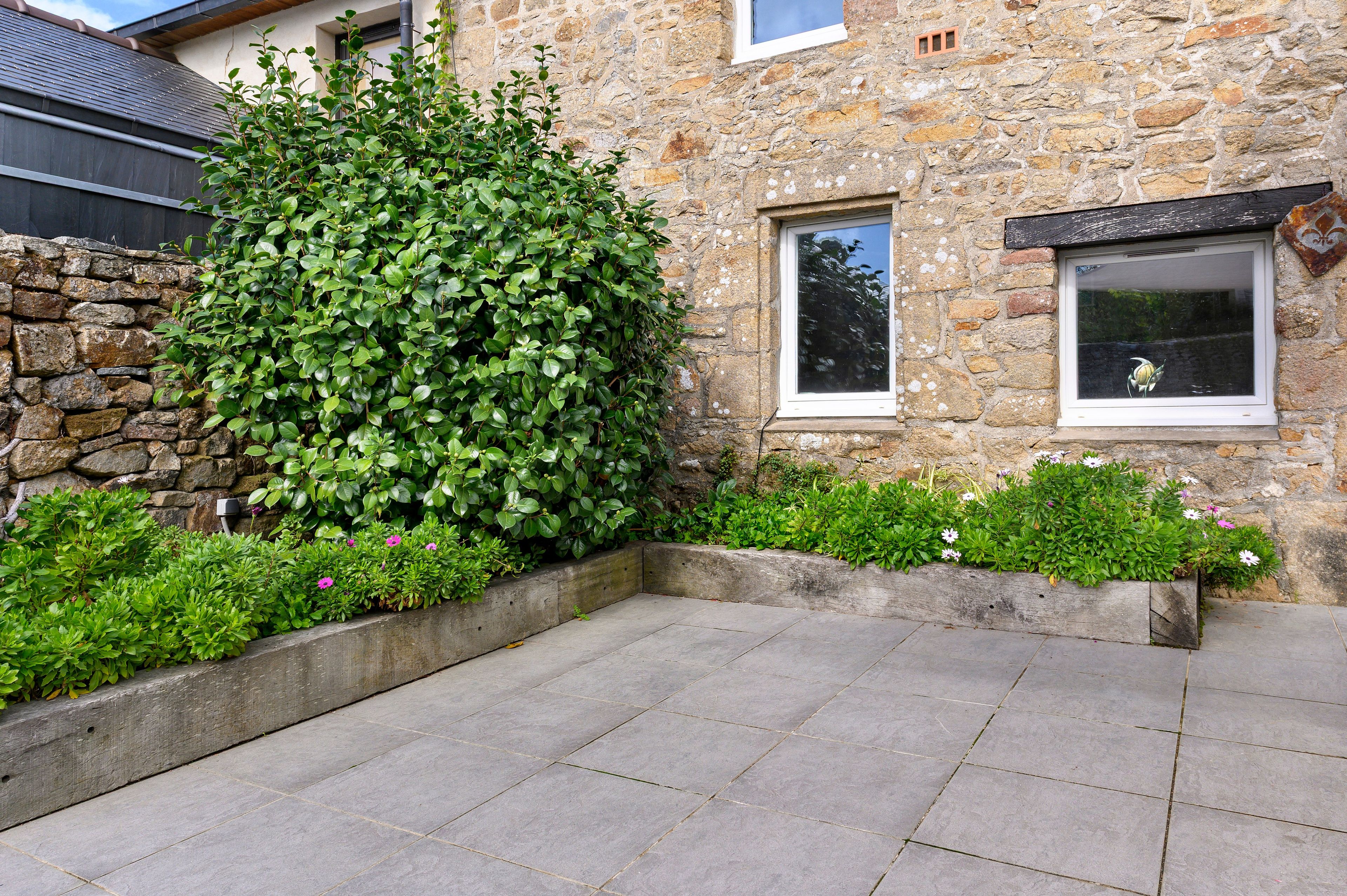 A stone building with a paved courtyard, a large hedge, and a small window with a decorative element.