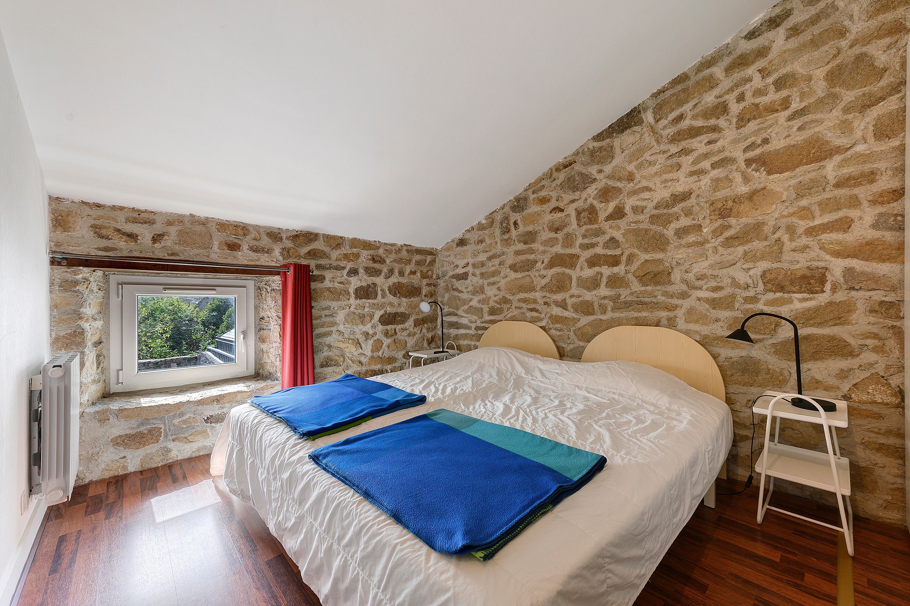 A bedroom with a stone wall, a window with a view, a bed with two pillows, and a bedside table with a lamp.