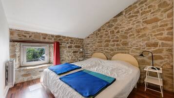 A bedroom with a stone wall, a window with a view, a bed with two pillows, and a bedside table with a lamp.
