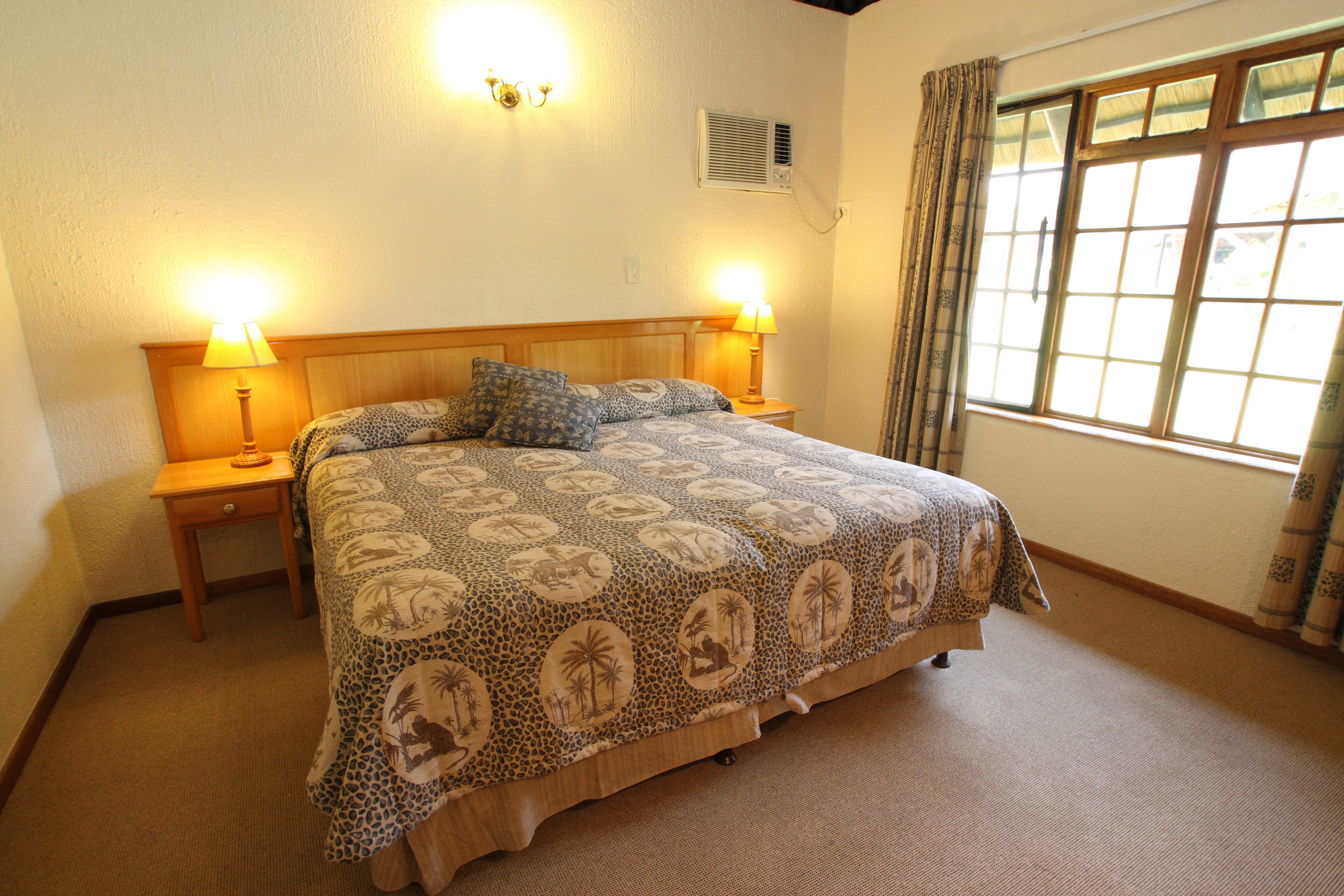 A bedroom with a bed, two bedside lamps, a window with curtains, and an air conditioning unit on the wall.