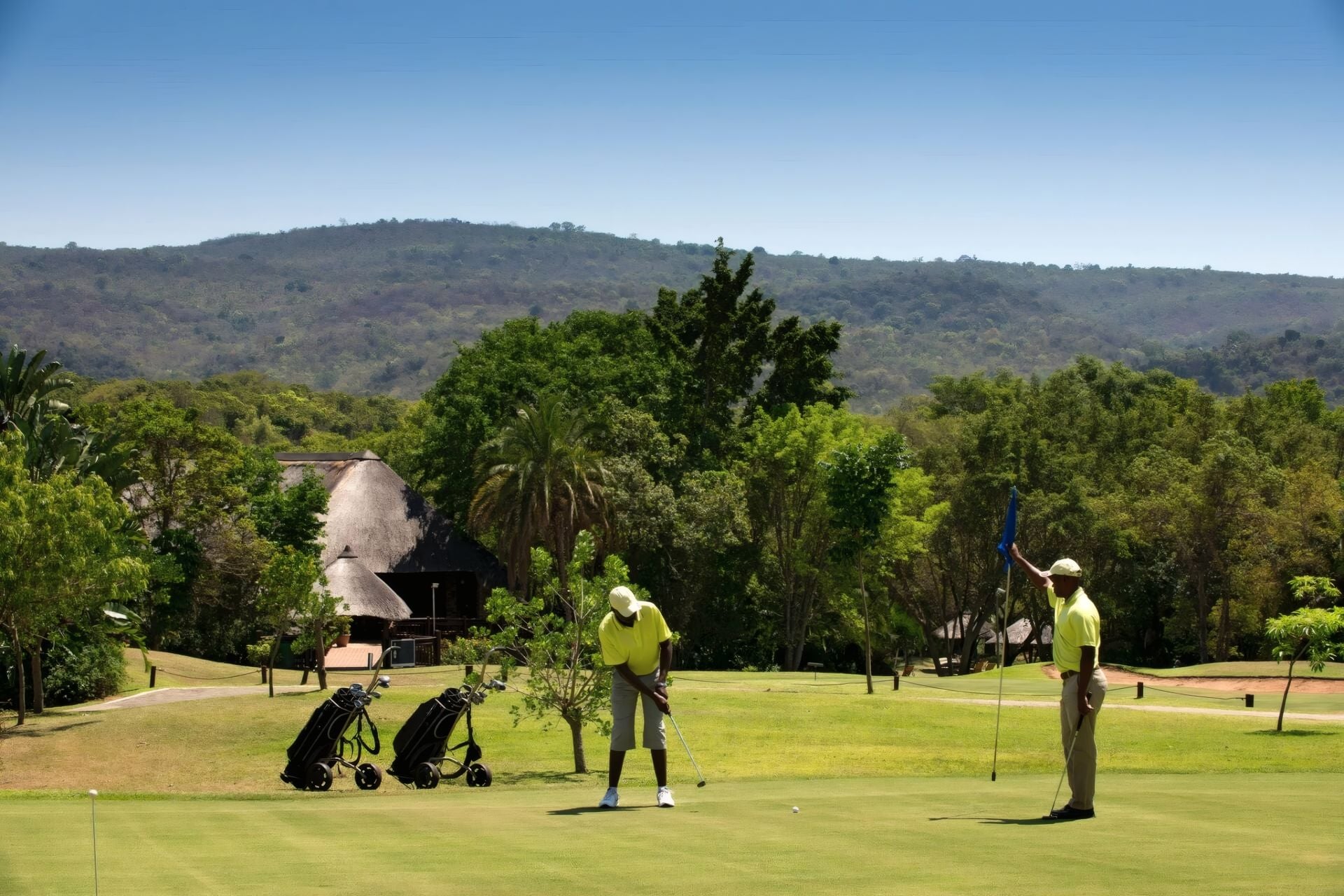 Un campo de golf con un edificio de techo de paja, bolsas de golf y jugadores.