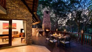 An outdoor dining area with a brick fireplace, surrounded by a wooden deck and overlooking a forested area.