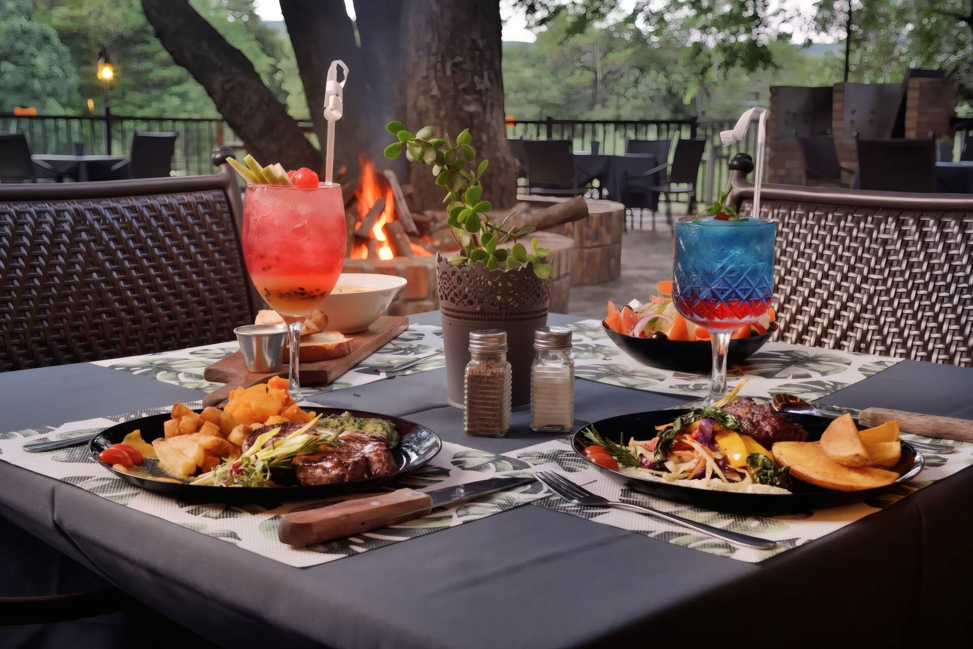 An outdoor dining area with a table set for two, featuring a variety of dishes, drinks, and a fire in the background.