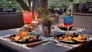 An outdoor dining area with a table set for two, featuring a variety of dishes, drinks, and a fire in the background.