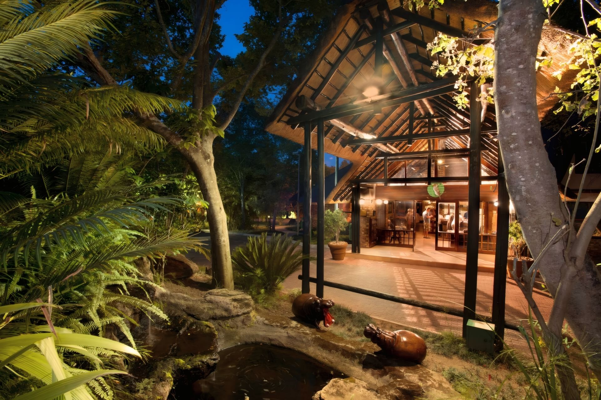 A building with a covered outdoor area, surrounded by tropical vegetation and a small water feature with hippo statues.