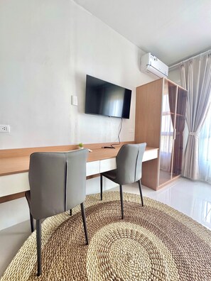 A modern living room with a flat-screen TV mounted on the wall, a wooden desk, two grey chairs, a woven rug, and a large window with curtains. - Royal Oceancrest Panglao 1 (Dauis)