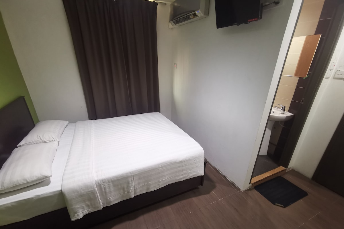 A single bed with a white mattress and pillows, a dark headboard, a brown curtain, a wall-mounted air conditioning unit, a television, and a small bathroom with a sink and mirror.