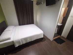 A single bed with a white mattress and pillows, a dark headboard, a brown curtain, a wall-mounted air conditioning unit, a television, and a small bathroom with a sink and mirror. - Red Tomato Hotel - self check in (Labuan)