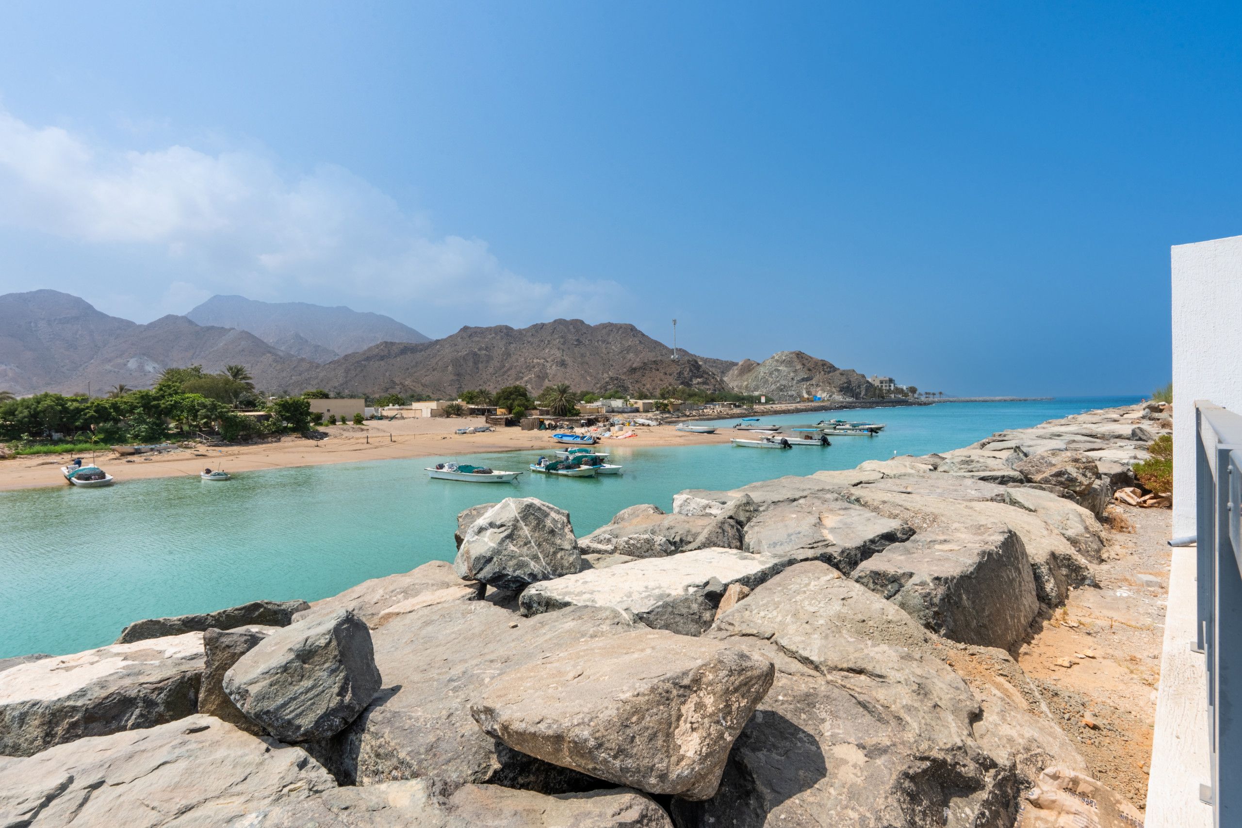A coastal landscape with rocky terrain, a sandy beach, and a clear blue sea. Boats are moored along the shore, and mountains are visible in the background.