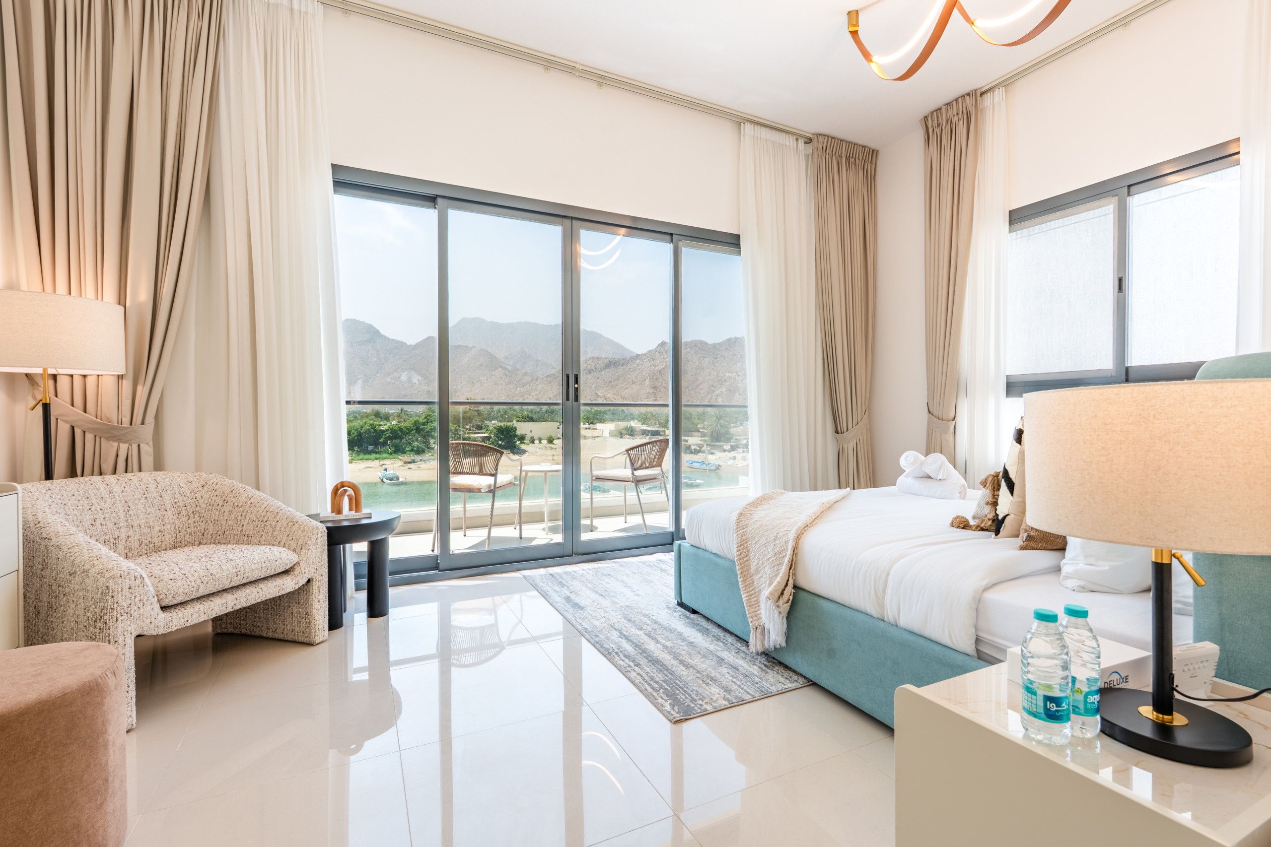 A hotel room with a bed, a sofa, a table with a lamp, a view of mountains and a pool.