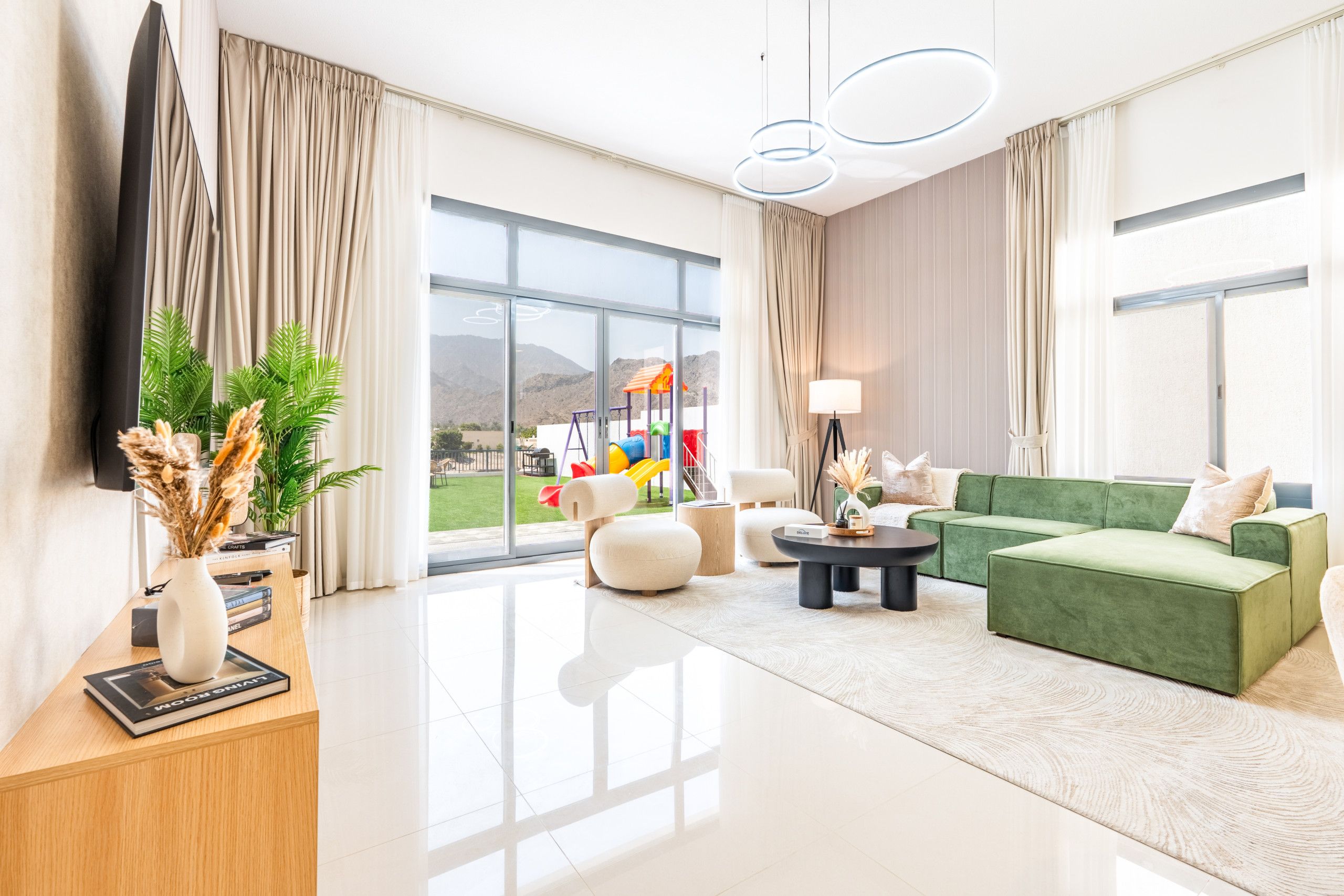 Una sala de estar moderna con un sofá verde, una mesa de centro y vista a un parque infantil a través de grandes ventanales.