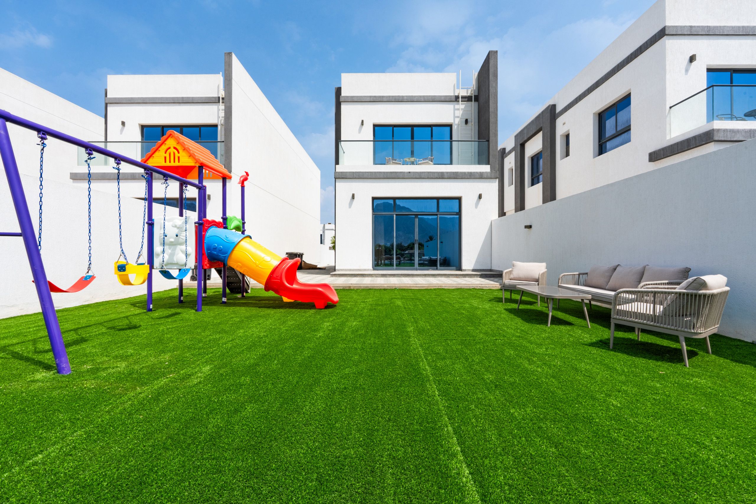Un área de juegos exterior moderna con un tobogán y columpios, contigua a un edificio con amplias ventanas y un balcón.