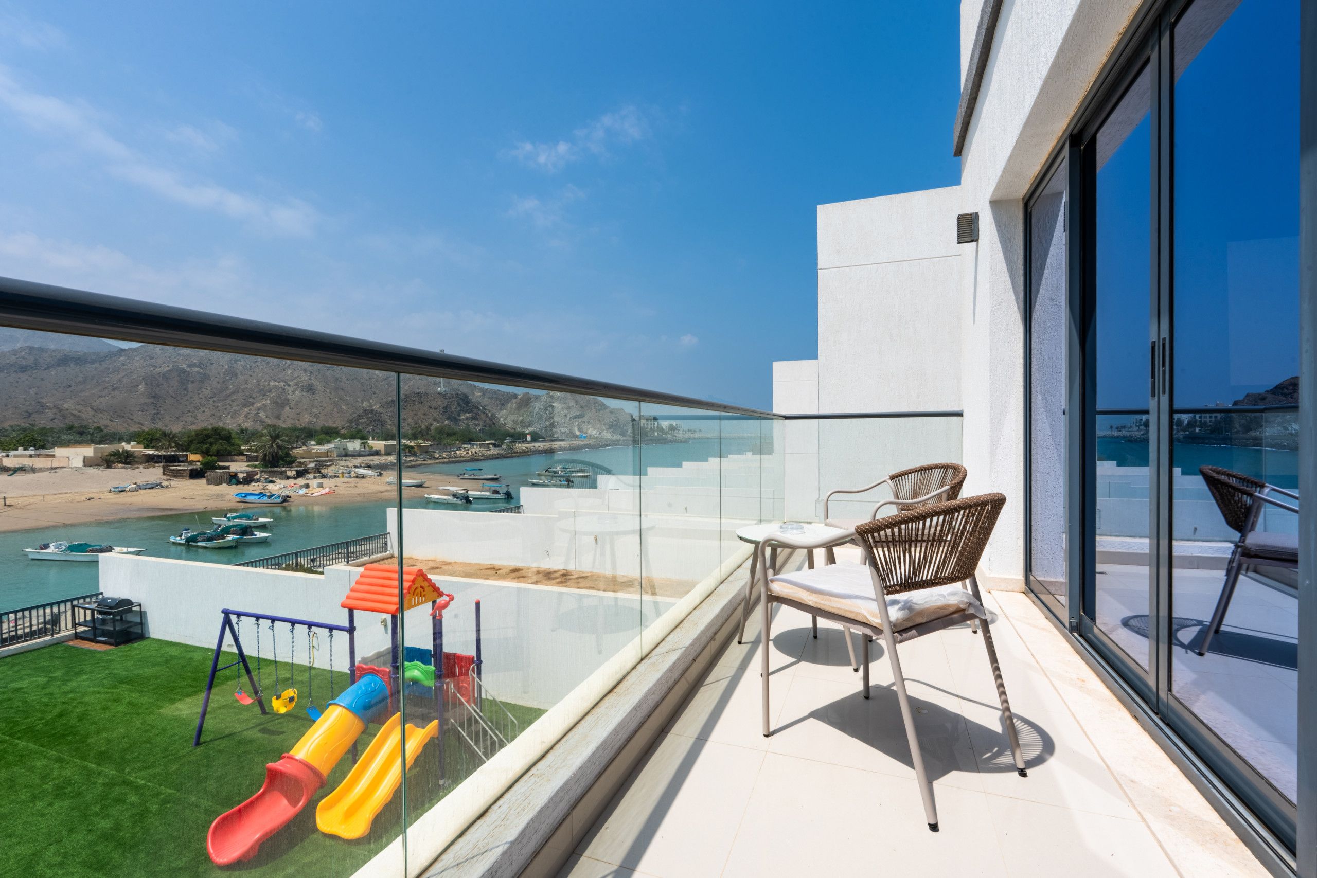 A balcony with a view of a beach, boats, and a playground.