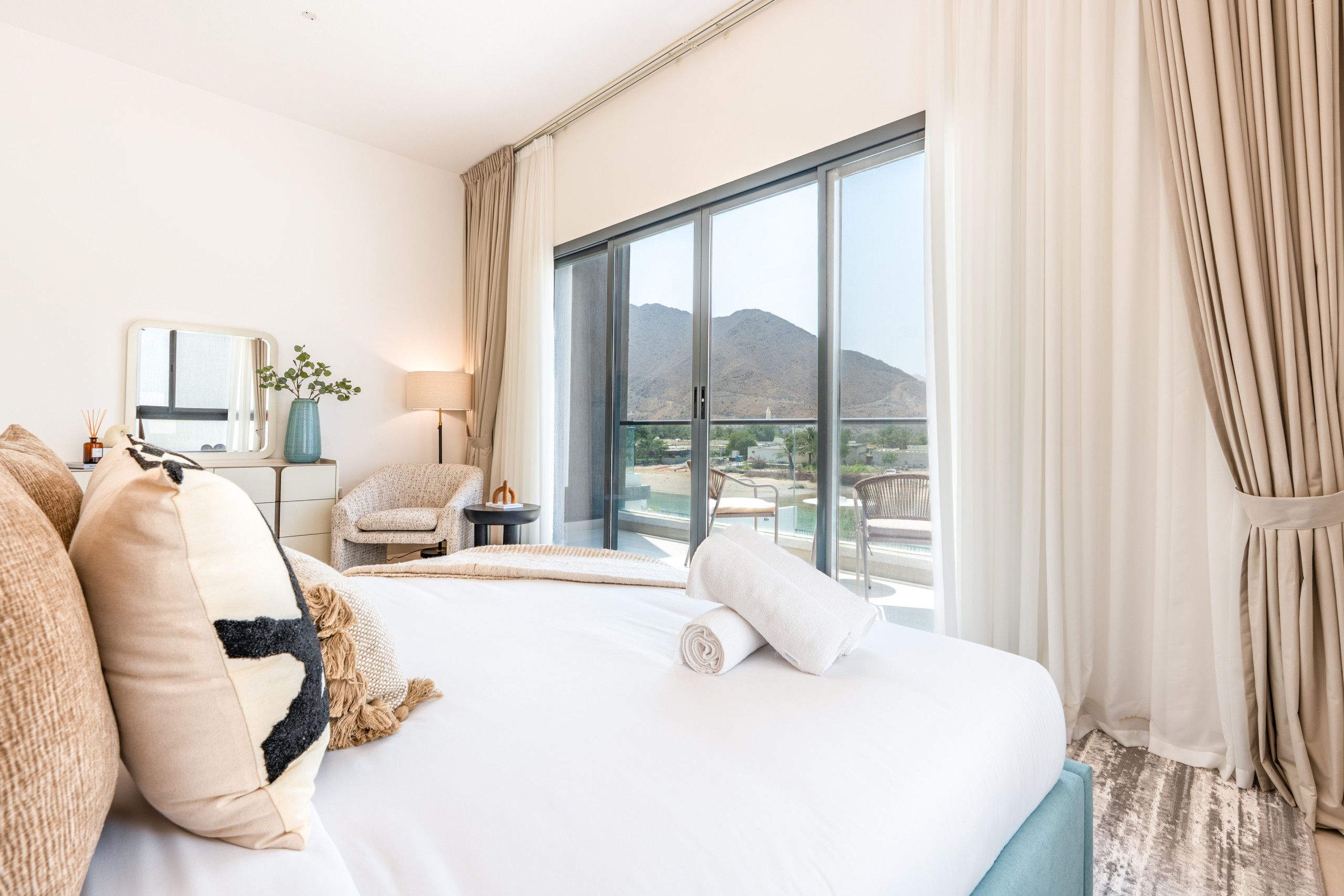A hotel room with a large bed, a chair, a side table, and a view of mountains through a large window.
