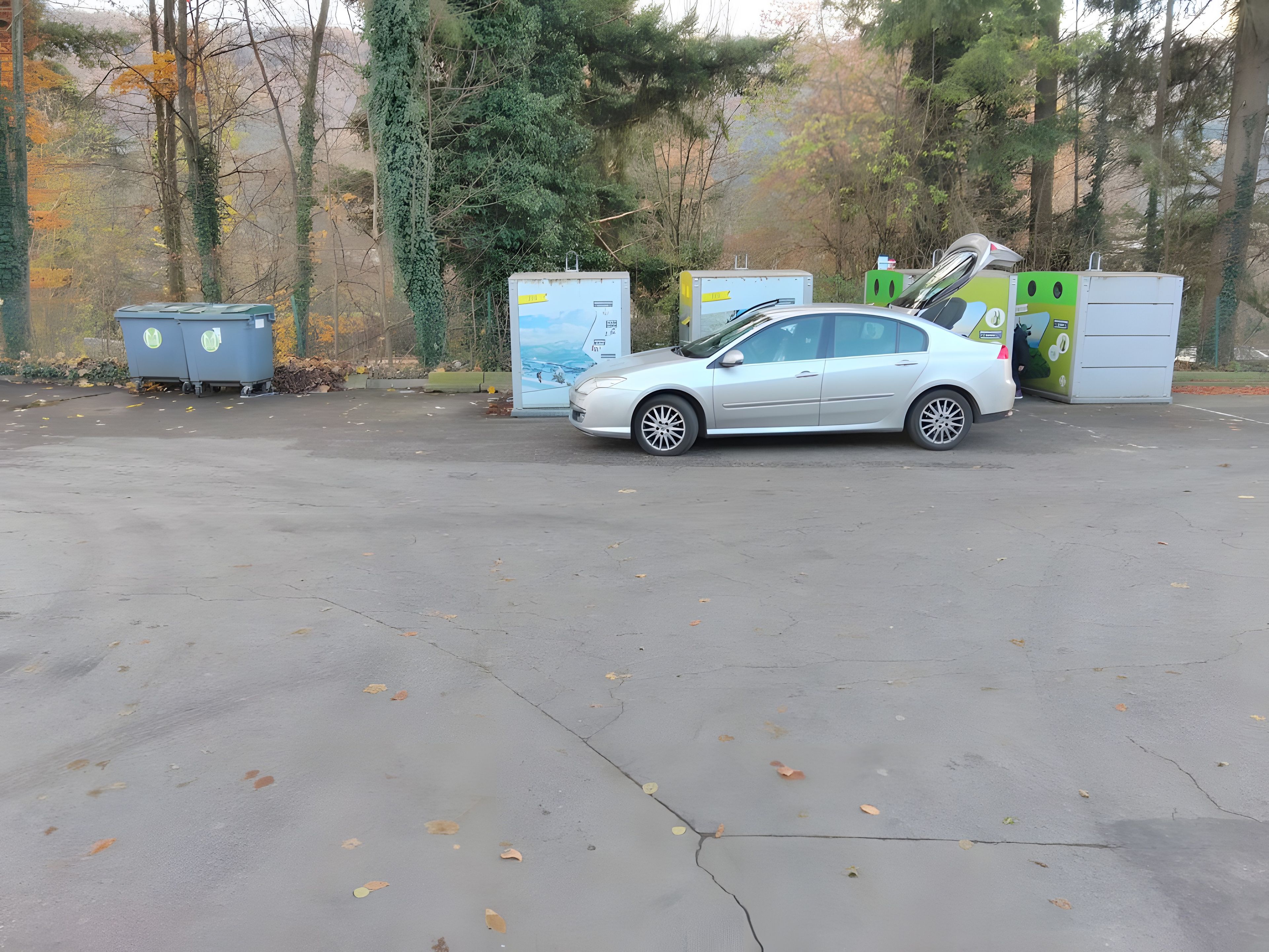 Ein Parkplatz mit einem weißen Auto, das neben Mülltonnen und einem grünen und weißen Recyclingbehälter steht.