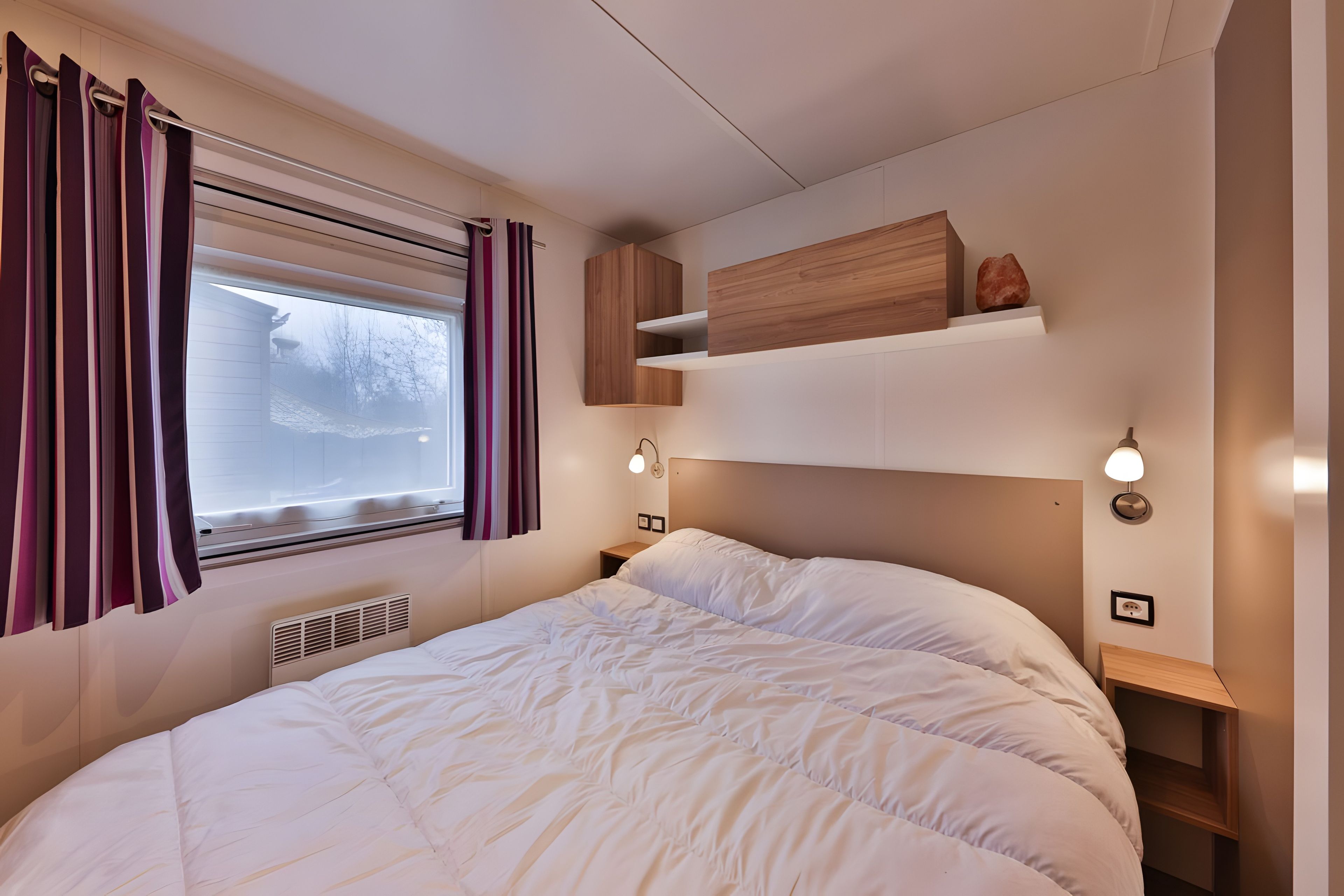 Quarto com cama, janela com cortinas, estante embutida, luminárias de cabeceira e aquecedor fixado na parede.