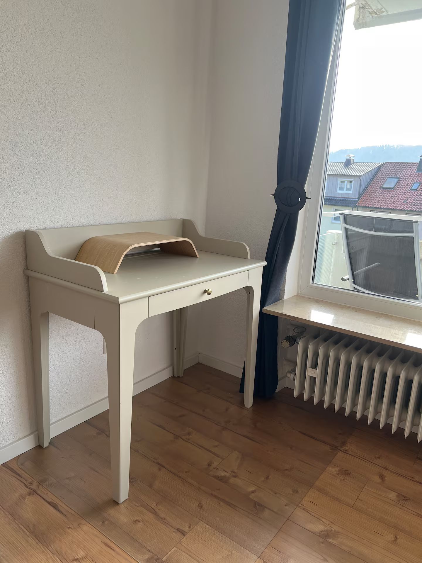Ein Zimmer mit einem weißen Schreibtisch, einem Heizkörper und einem Fenster mit Blick auf Gebäude.