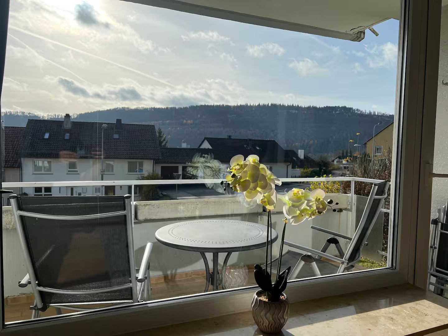Ein Balkon mit Tisch und Stühlen, einem Blumentopf und Blick auf Häuser und Hügel.
