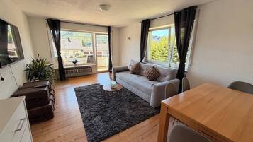 Ein möbliertes Wohnzimmer mit einer Couch, einem Esstisch, einem kleinen Teppich und Blick durch große Fenster.