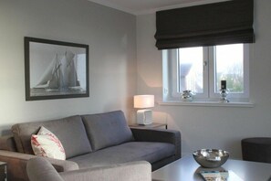 A modern living room with a grey sofa, a framed picture of a sailboat, a window with a dark valance, a bedside table with a lamp, and a table with a bowl. - Holiday apartment in Sylt with 2 bedrooms (Sylt \/ OT Westerland)