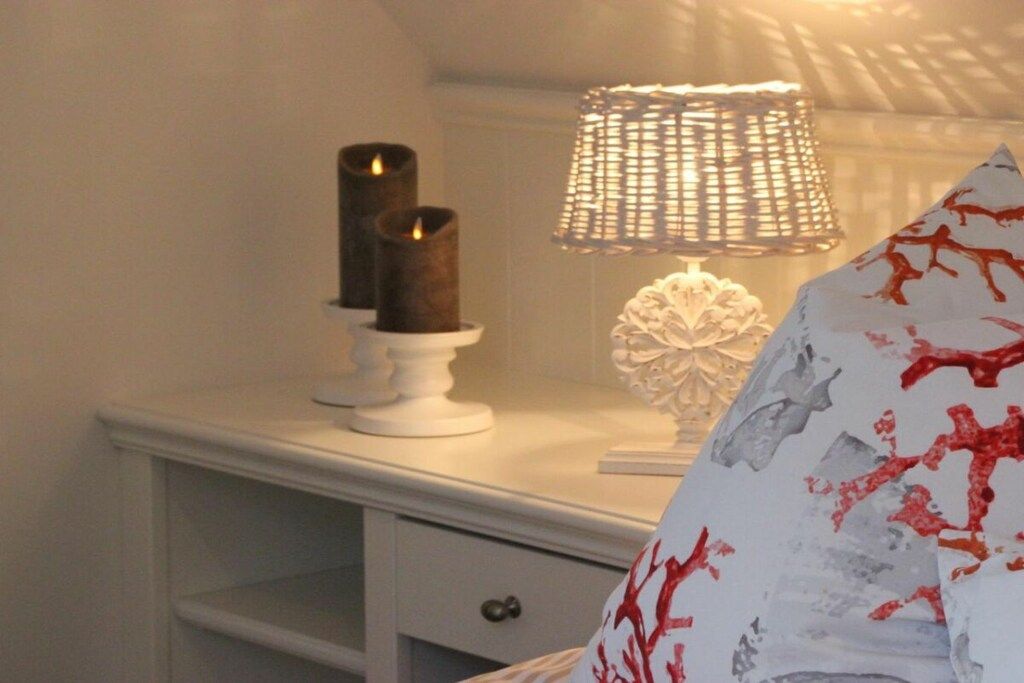 A white bedside table with a white lamp, two candles, and a decorative white object.