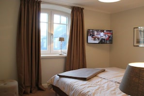 A hotel room with a bed, curtains, a television on the wall, and a lamp. - Holiday apartment in Sylt with 2 bedrooms (Sylt \/ OT Westerland)