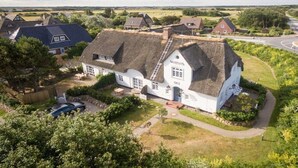 Une maison blanche traditionnelle, dotée d’un toit de chaume, entourée de verdure et bordée par une route.