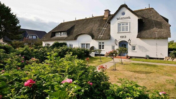 Un bâtiment traditionnel, de couleur blanche, avec un toit de chaume, portant l’inscription « WITHHUS 1913 », et entouré d’une végétation luxuriante et de plantes en fleurs.