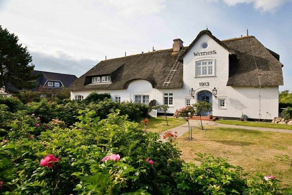 A traditional white building with a thatched roof, labeled 'WITHHUS 1913,' surrounded by lush greenery and flowering plants. - Witthüs 15 - stylish living under a thatched roof (Wenningstedt)
