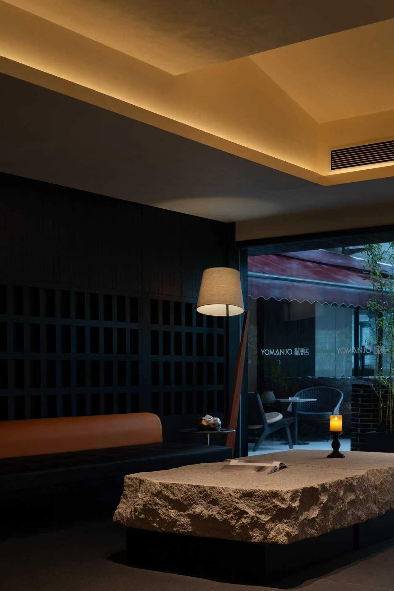 Un interior moderno con una recepción de textura de piedra, una lámpara y una zona de estar con un banco y una mesa.