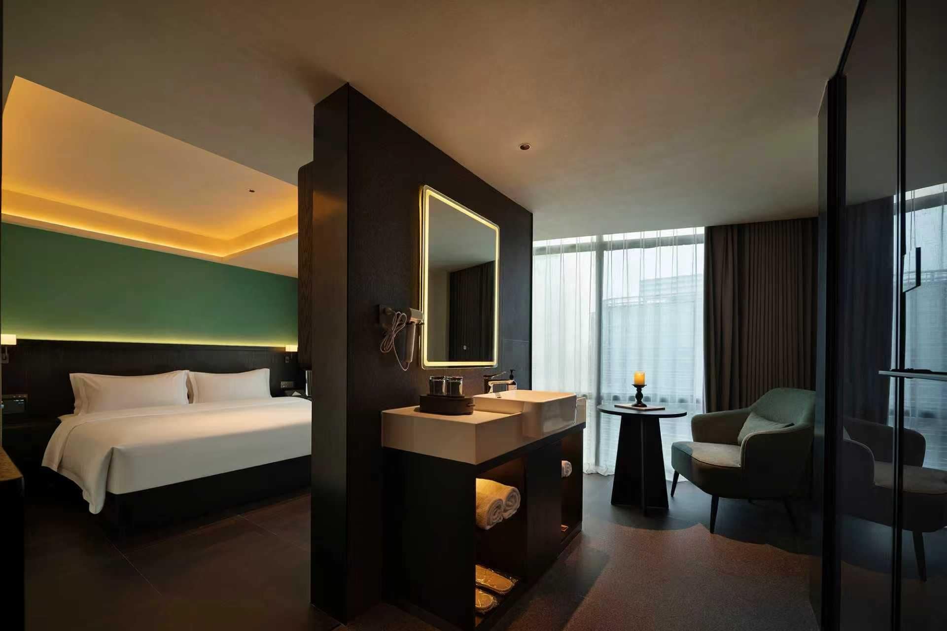 A hotel room with a bed, a large mirror, a vanity with a sink, a chair, and a view of the city through large windows.