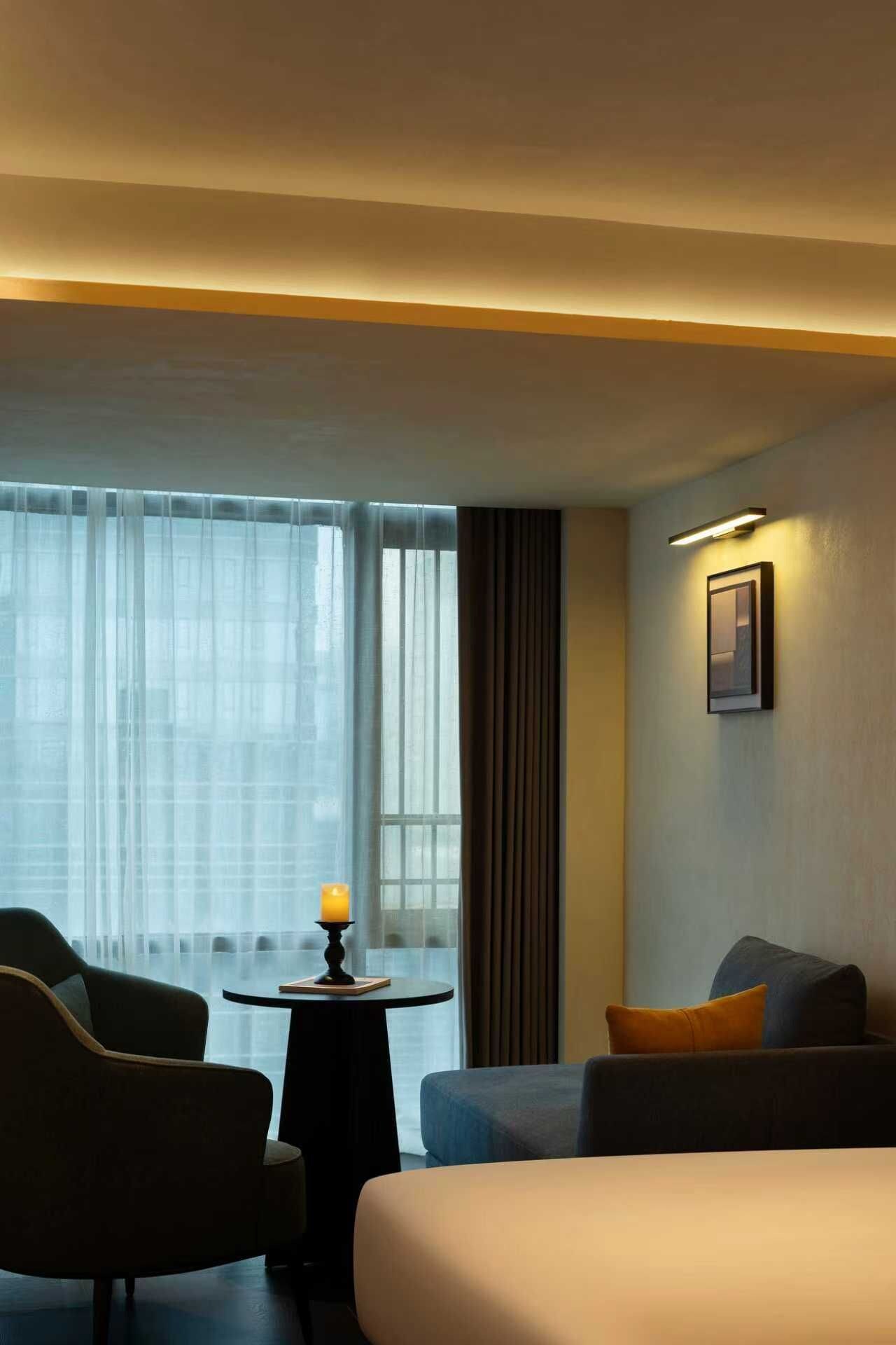 Une chambre d’hôtel comprenant un lit, deux fauteuils, une petite table avec une bougie, une fenêtre avec des rideaux et un luminaire fixé au mur.