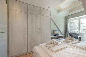 Quarto com um guarda-roupa espaçoso, cama com toalhas, televisão sobre um suporte e janela com vista para prédios.