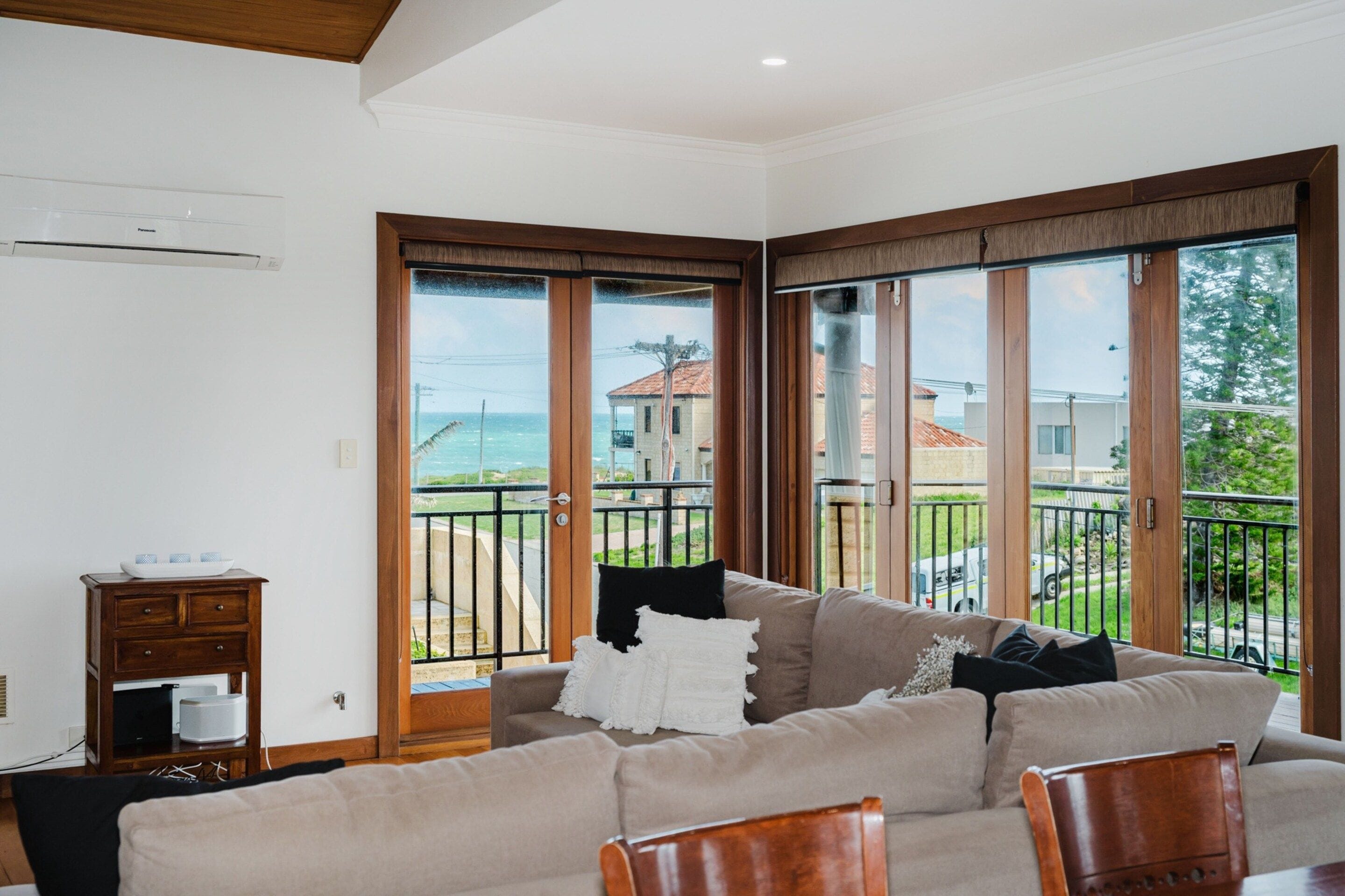 A living room with a beige sofa, wooden furniture, and large windows with a view of the sea and buildings.