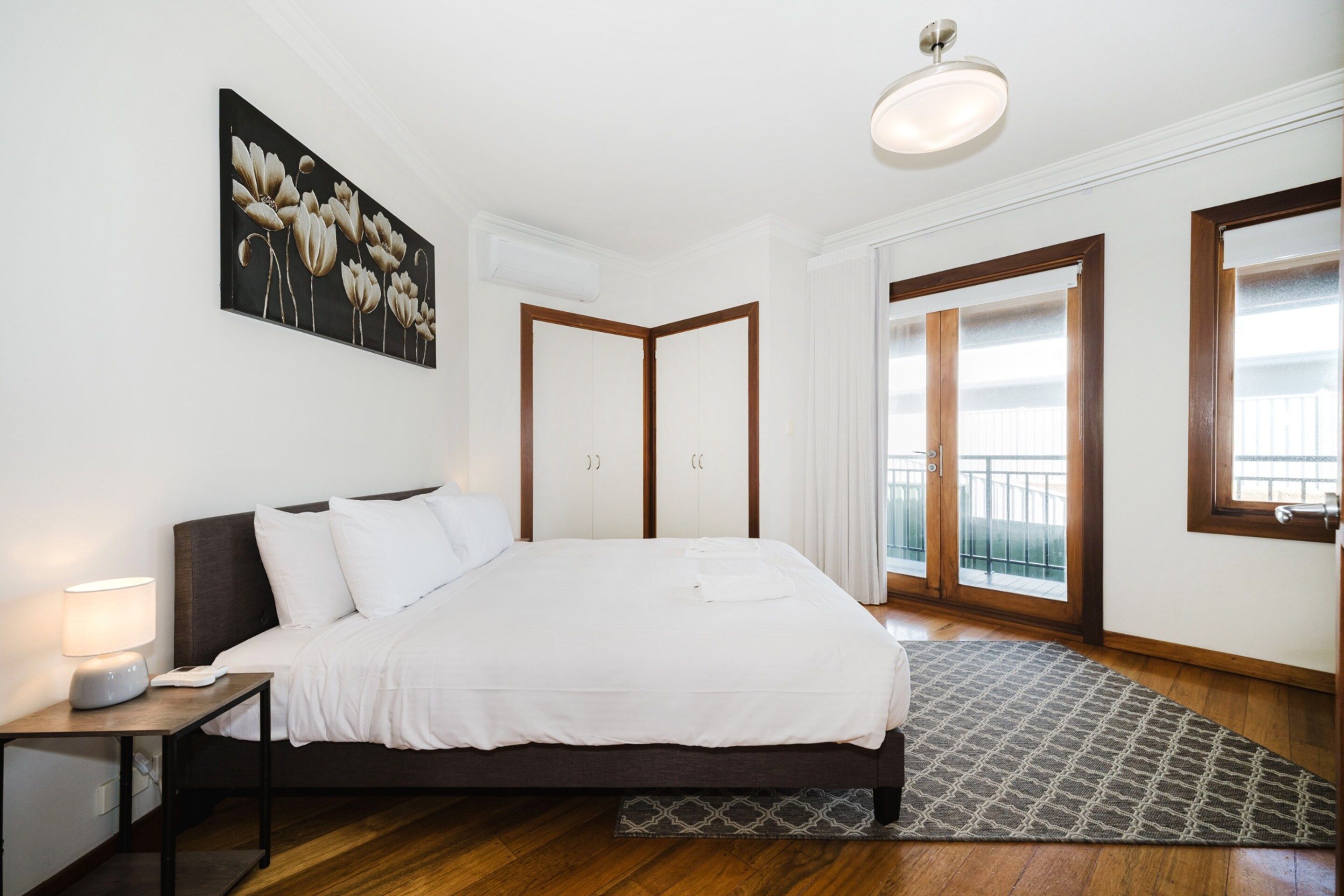 A bedroom with a bed, bedside table, lamp, artwork, and a view of the outside.
