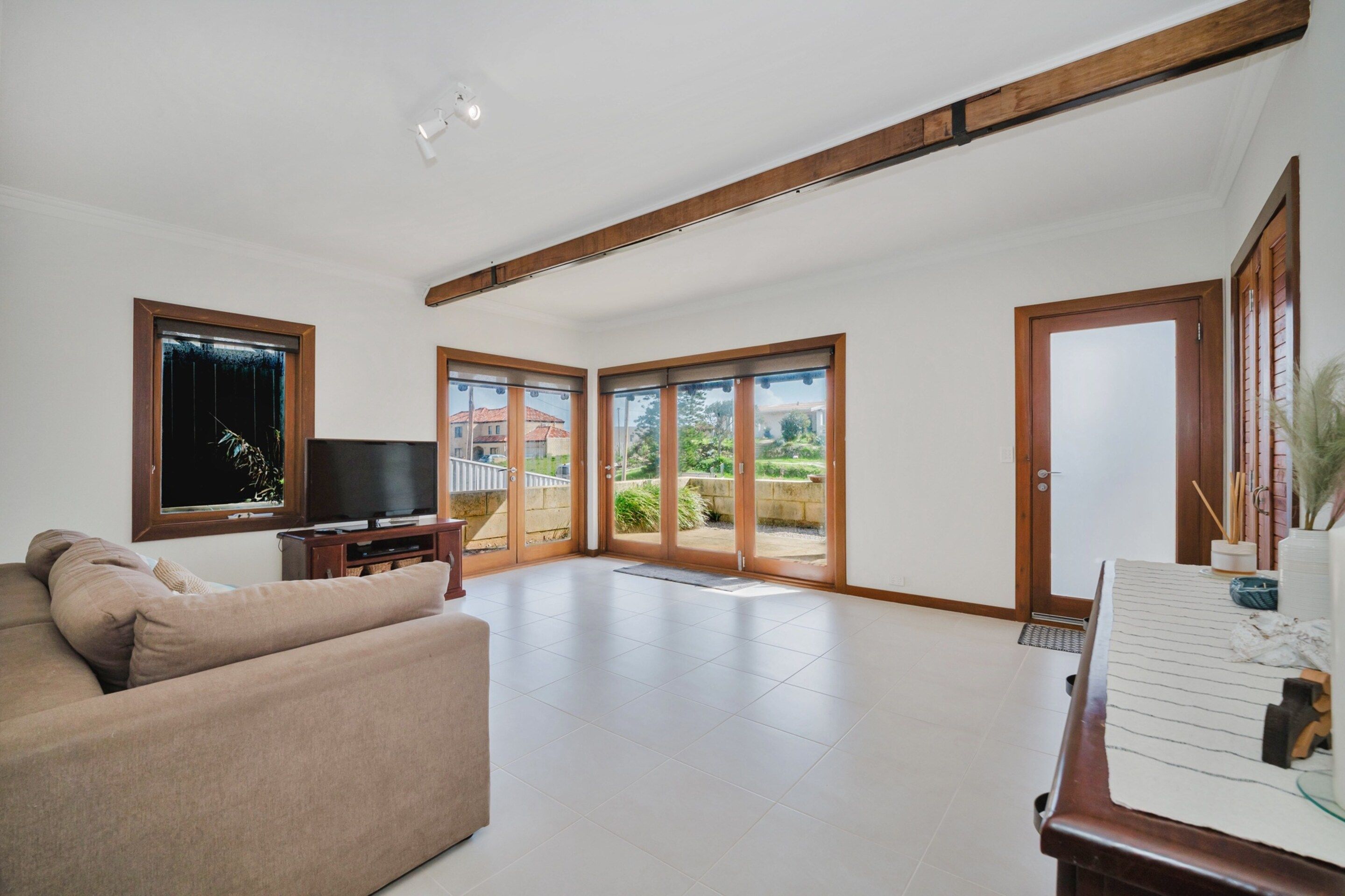 A spacious living room with a large window, a flat-screen TV, a sofa, and a wooden ceiling beam.