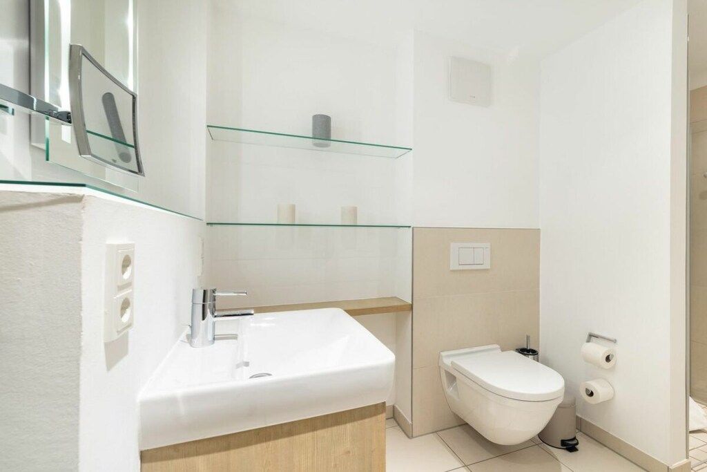 A modern bathroom with a white sink, a toilet, and glass shelves above.