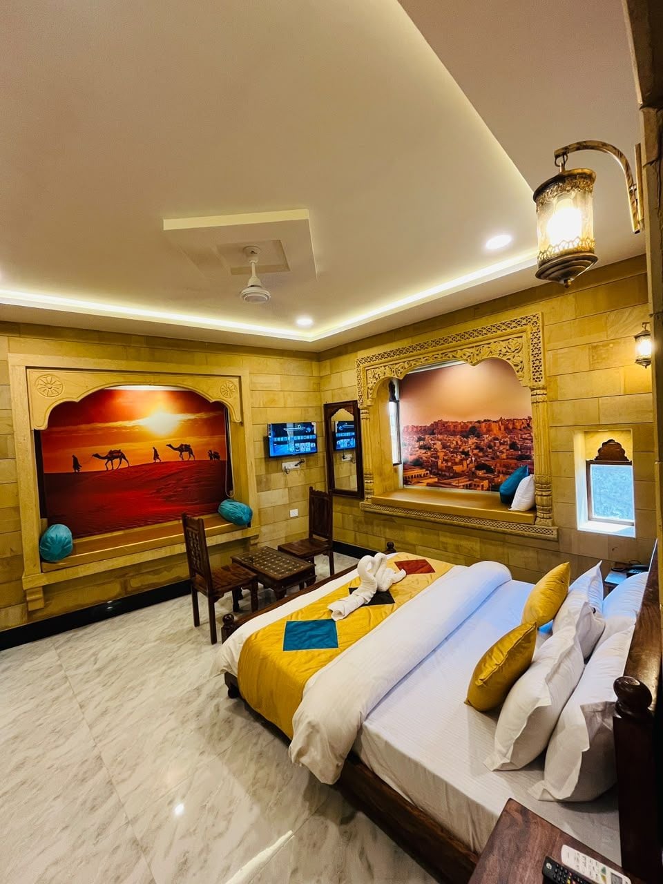A hotel room with a bed, a television, a wall-mounted TV, and a decorative wall with a desert scene.