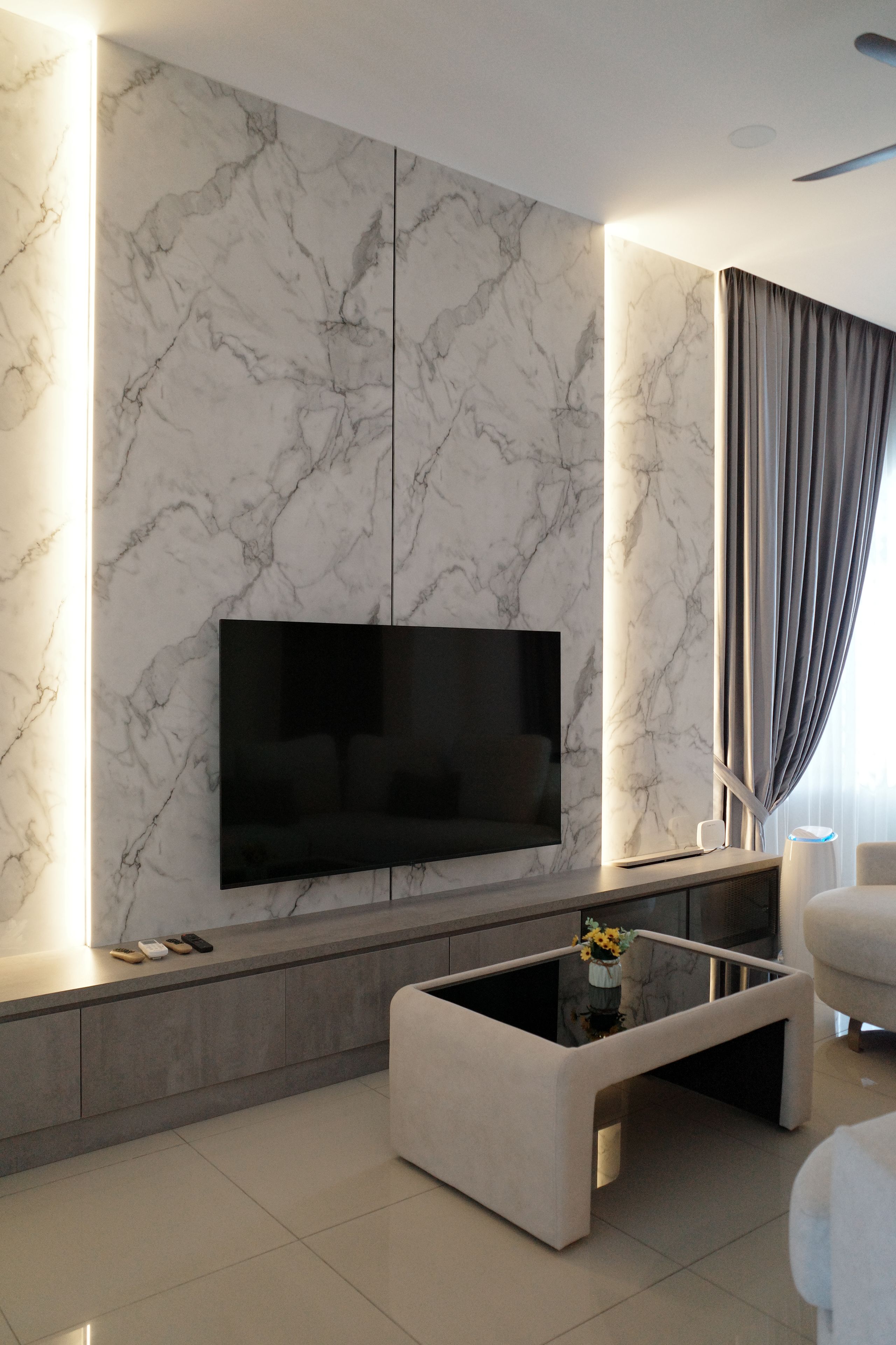 Ein modernes Wohnzimmer mit einem großen Flachbildfernseher an einer Marmorwand, einem Glastisch und einem Sofa.