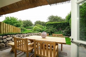 An outdoor patio with a wooden table and chairs, a stone wall, and a thatched roof. - Holiday home in Sylt with swimming pool (Sylt)